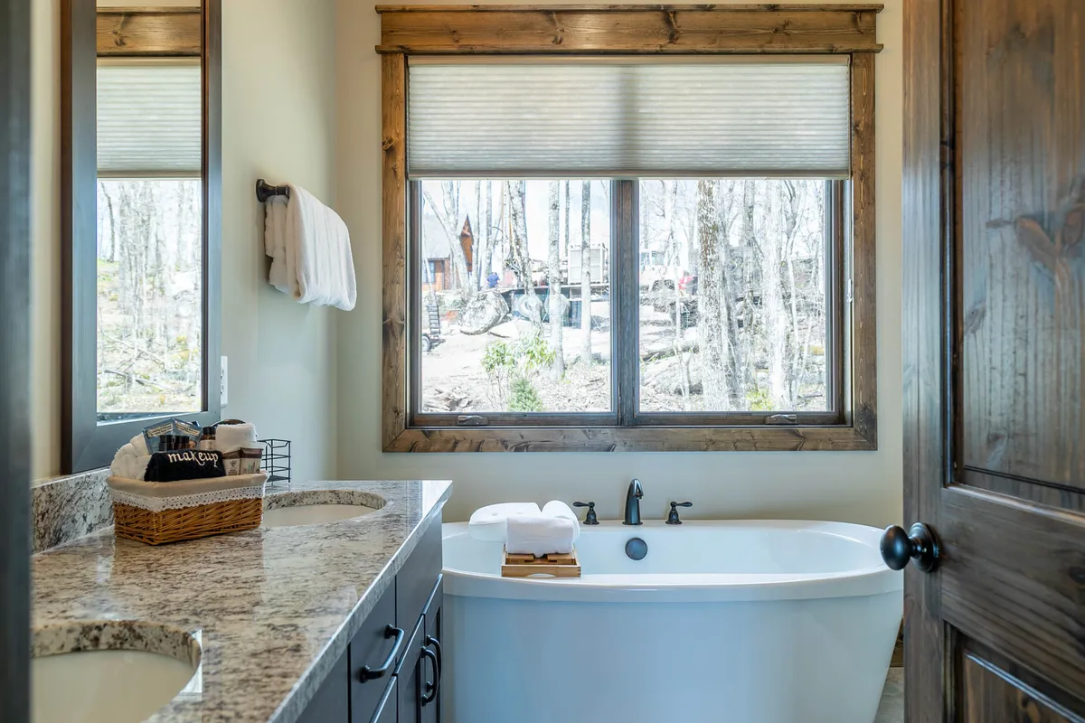 A serene bathroom featuring a freestanding bathtub positioned beside a large window that lets in natural light, complemented by a granite countertop and wooden accents. This space exudes tranquility and luxury, ideal for restful self-care routines.