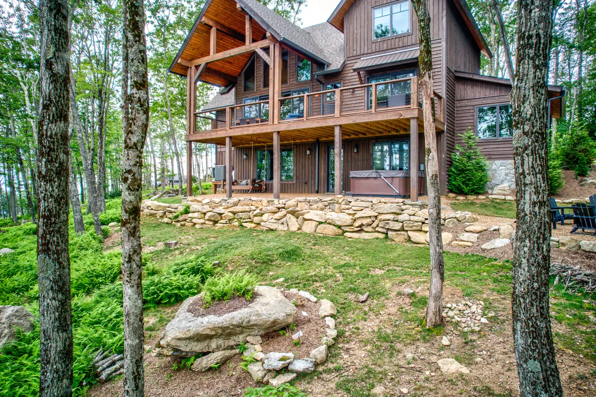 This image showcases a spacious wooden deck bordered by a stone wall and surrounded by lush greenery. Large windows connect the outdoor area to the interior of the home, emphasizing the seamless transition between indoor comfort and the natural world outside. The deck looks inviting for lounging or spending time with pets in a beautiful mountain setting.
