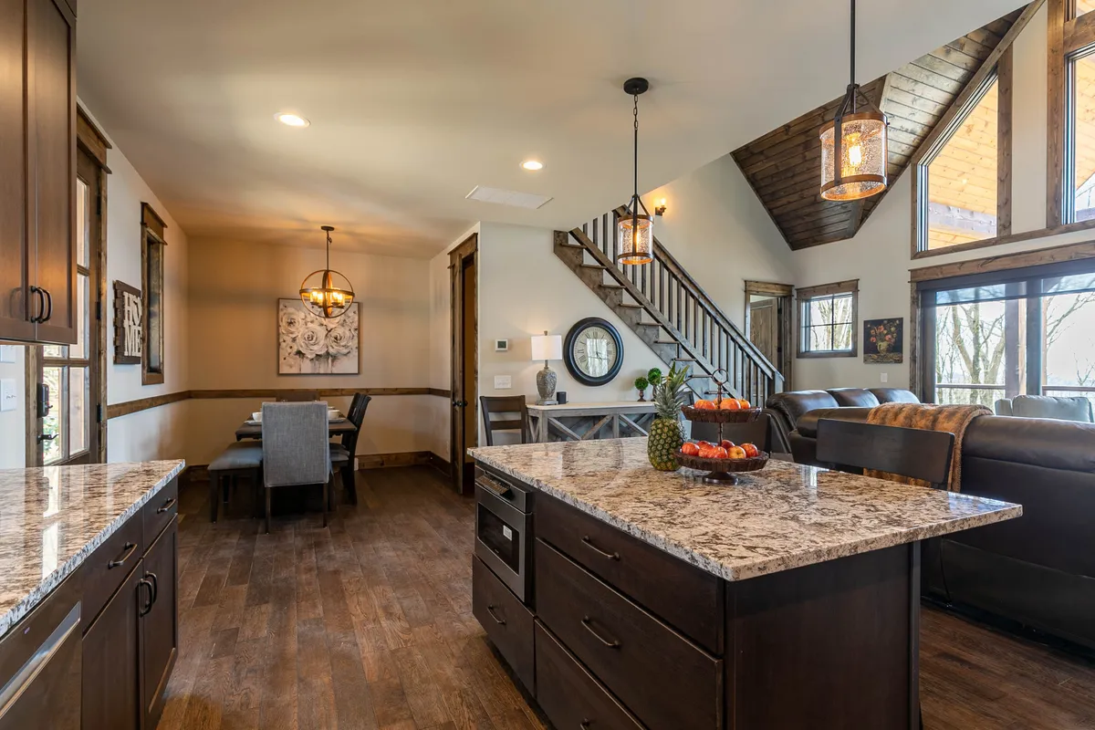 A warmly lit, open-concept interior featuring a granite countertop kitchen with stainless steel appliances. The space flows into a dining area with a wooden table and chairs and a cozy seating area, all bordered by large windows that reveal scenic mountain views. A wooden staircase and comfortable living arrangements create an inviting communal atmosphere.