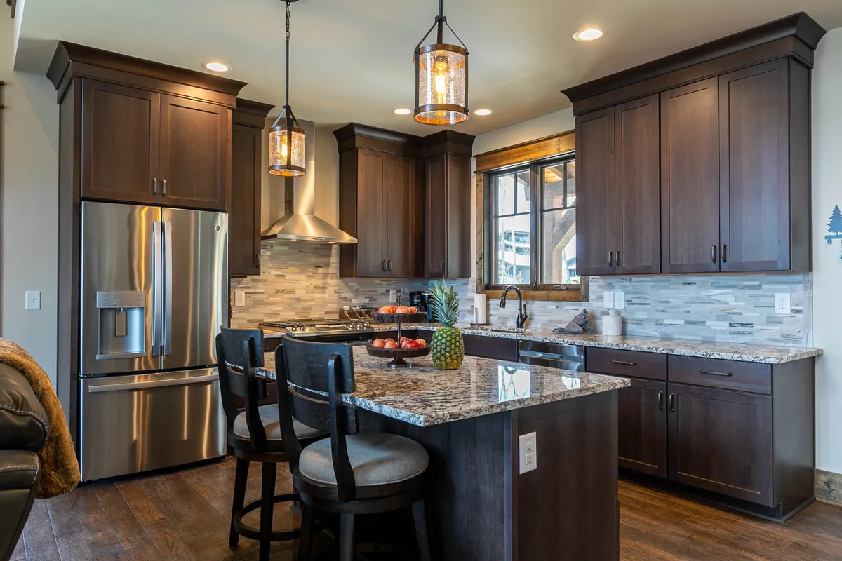 A modern yet cozy kitchen with a granite countertop island, stainless steel appliances, pendant lighting, and wooden cabinetry. The kitchen is open to surrounding spaces, inviting both culinary creativity and social interaction. A window offers additional natural light.