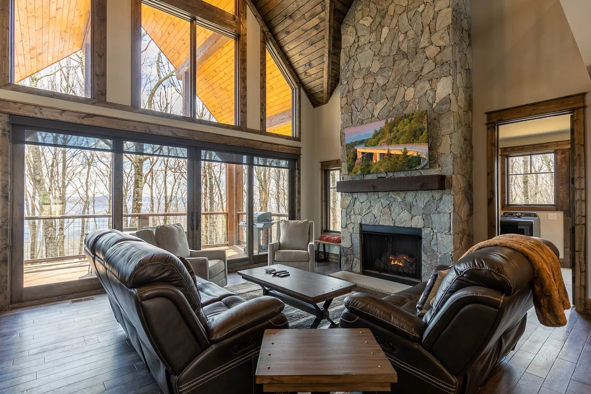 A welcoming living room scene highlighted by a stone gas log fireplace flanked by large windows. Leather sofas and wooden beams add to the cozy ambiance, while sunlight pours in, emphasizing the connection to the mountain landscape beyond.
