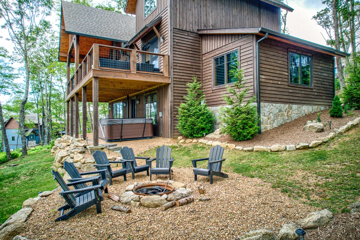 A spacious outdoor living area featuring a cozy fire pit surrounded by Adirondack chairs, a deck with an inviting hot tub, and natural stone landscaping. The deck is nestled among tall trees, creating a serene, private mountain setting ideal for fireside gatherings and relaxation.