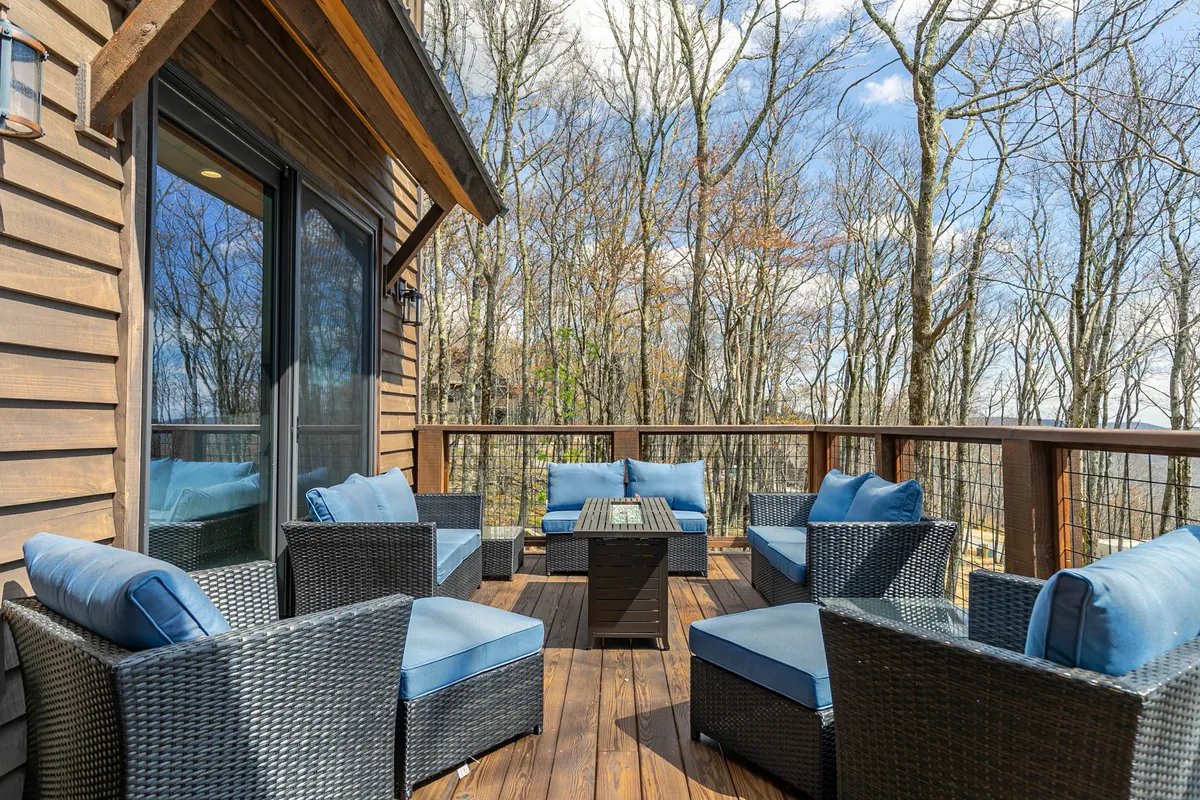 An inviting outdoor seating area on a wooden deck, surrounded by trees and accessible through glass doors. The setup offers a tranquil space for relaxing, reading, or spending time with a pet in the fresh mountain air.