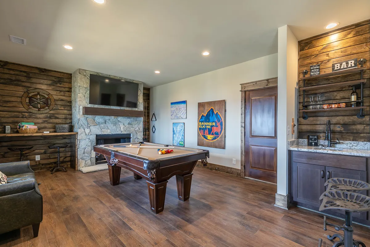 A well-appointed entertainment lounge featuring a pool table as the central attraction, with a stylish bar area to the side, a stone gas log fireplace, and artwork on the walls. Comfortable seating invites guests to relax and socialize, while the warm lighting creates a cozy, inviting atmosphere.
