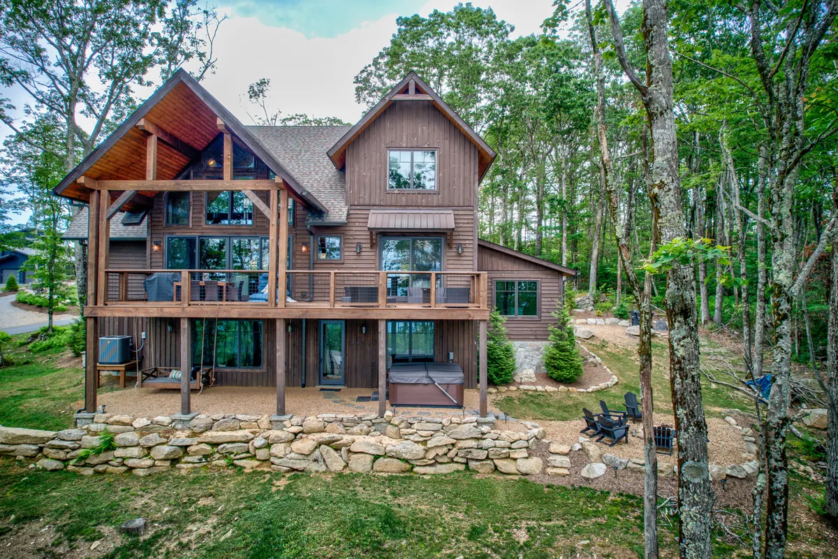 A wooden deck with comfortable outdoor seating, large windows connecting to the interior, a stone wall, and surrounding forest. The space is perfect for gatherings, outdoor meals, or simply enjoying the mountain air day or night.