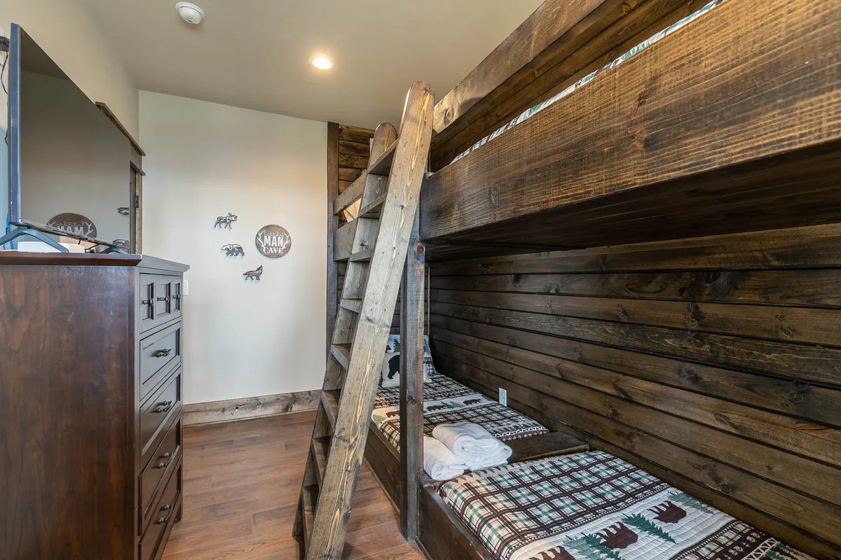 A playful bunk room featuring wooden bunk beds with plaid bedding, a sturdy wooden ladder, and a dresser. The warm color palette and cozy setup make this an ideal space for kids or multiple guests, inspiring sleepovers and late-night storytelling.