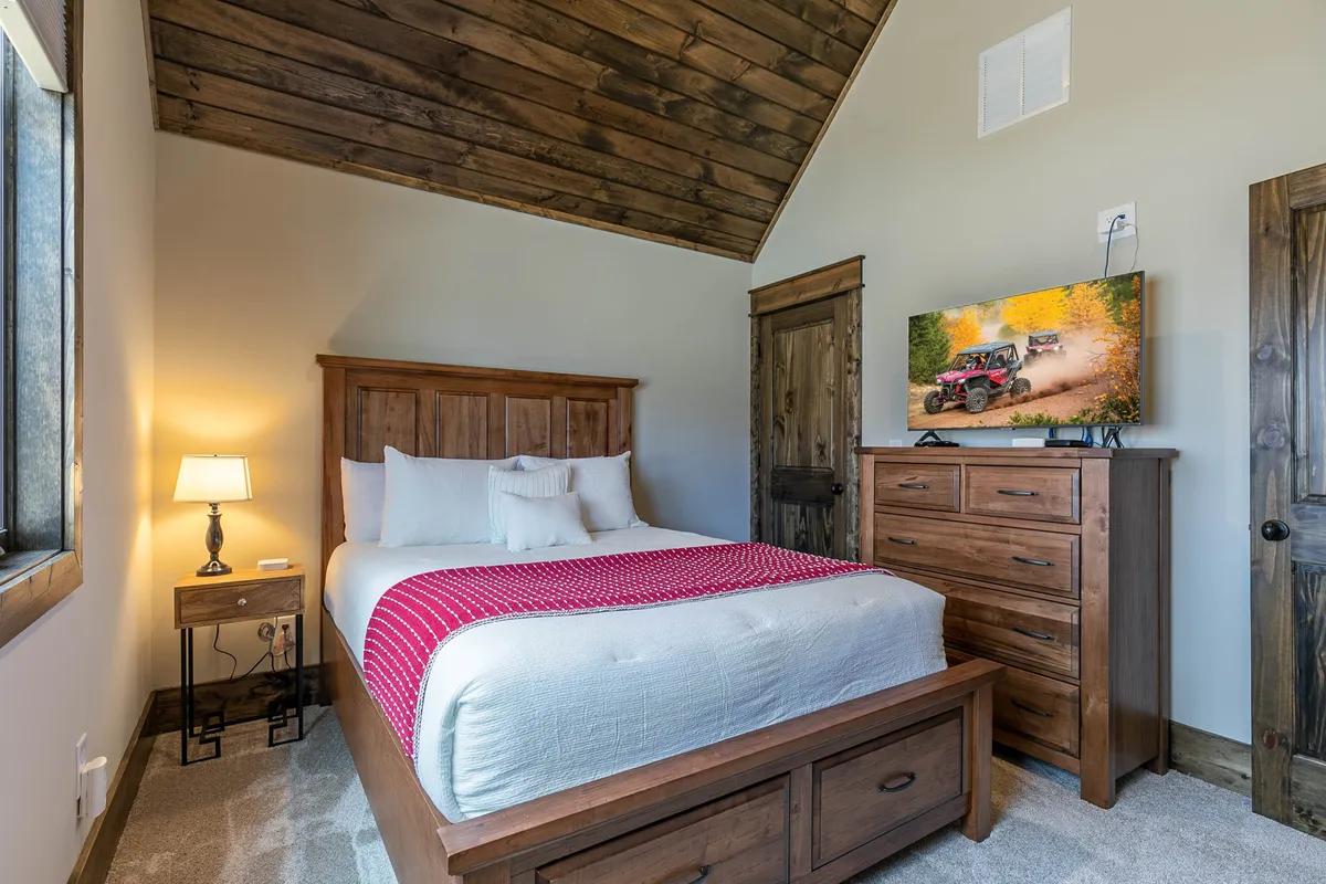 A cozy bedroom with a large bed, a nightstand and lamp for ambient lighting, and a dresser topped with a TV. The inviting layout offers both comfort and entertainment, reflecting a space made for relaxation and personal downtime.