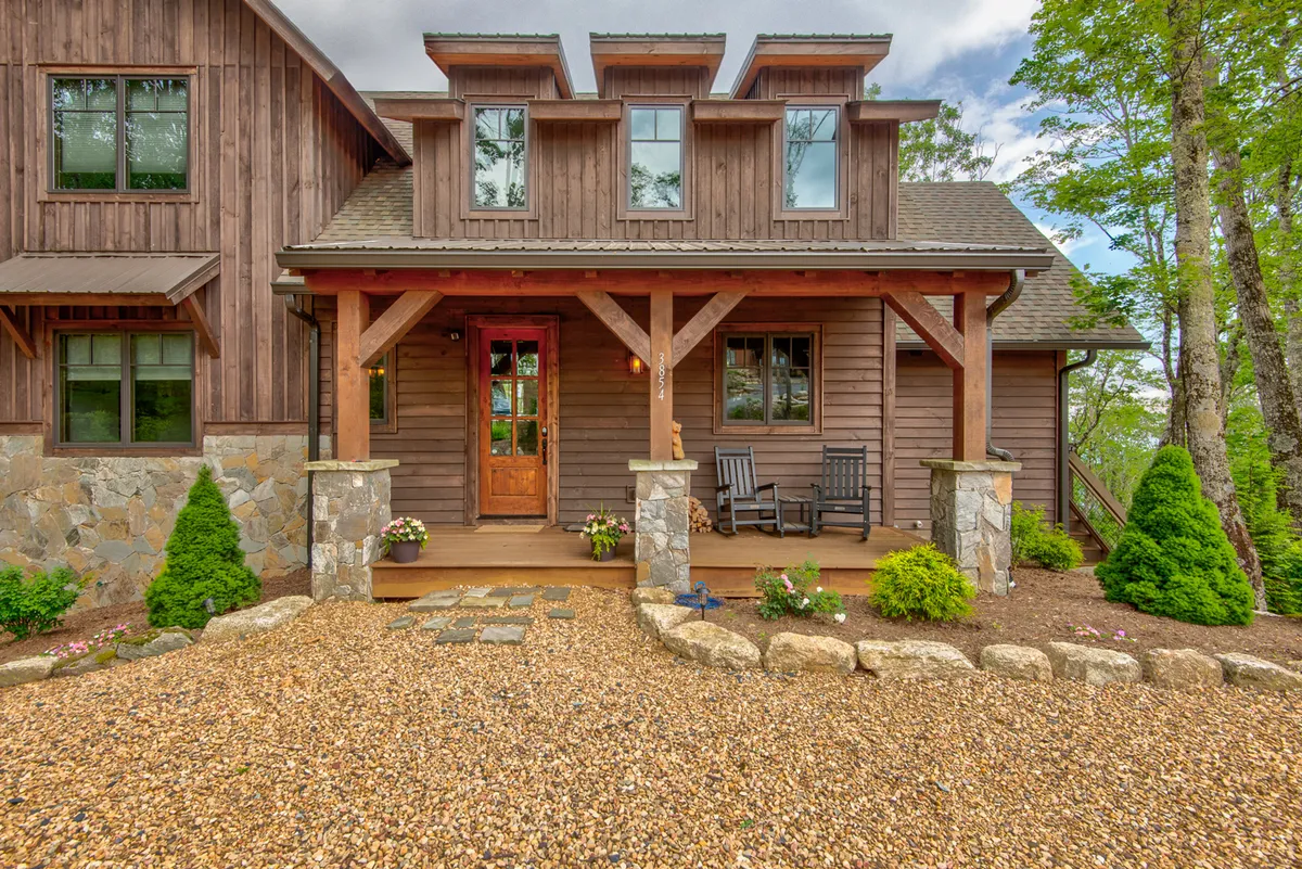 The front door of the lodge is shown with classic rocking chairs on the porch, a stone pathway, and lush garden landscaping. The setting exudes tranquility, ideal for enjoying sunsets or festive seasonal decorations, reinforcing the lodge as a home base for all occasions.